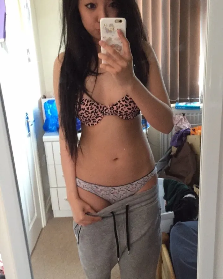 Femme trans asiatique aux cheveux longs noirs, prenant un selfie dans un miroir, en sous-vêtements léopard, avec un sourire discret.