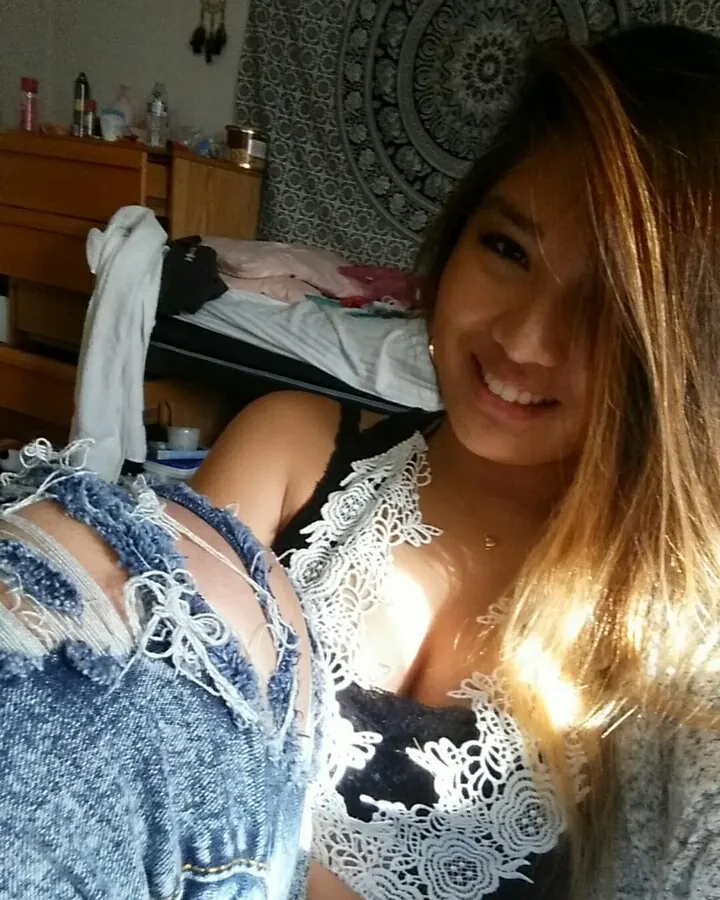 Femme trans latino aux cheveux bruns clairs, souriante, en haut noir à dentelle blanche et jeans déchirés, dans une chambre avec motif mandala au fond.