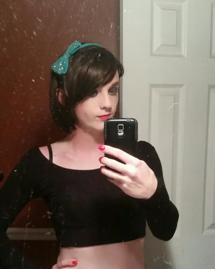 Femme trans européenne aux cheveux châtains courts, en haut noir et jeans, prenant un selfie dans un miroir, avec des ongles rouges et un sourire discret.