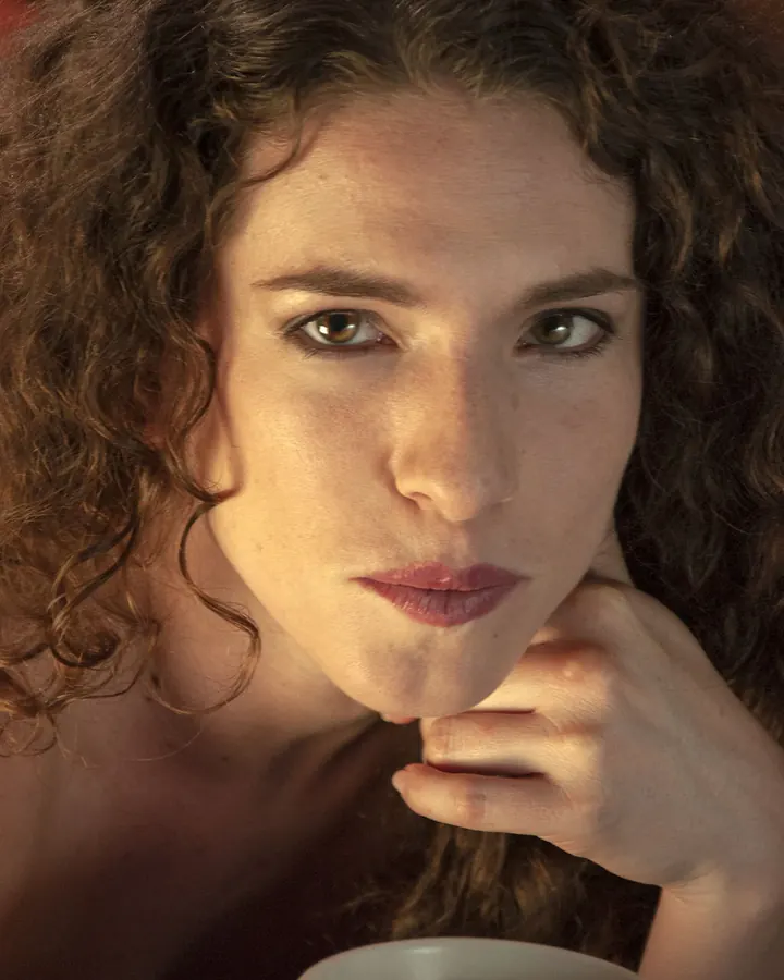 Femme trans aux cheveux bruns, portant un haut noir, regardant l'objectif dans un cadre intérieur.
