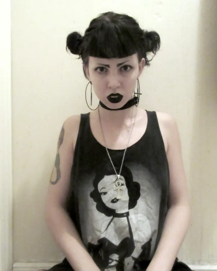 Femme trans latine aux cheveux bruns-noirs, en haut noir, posant devant un fond blanc, avec des lunettes noires et des piercings visibles
