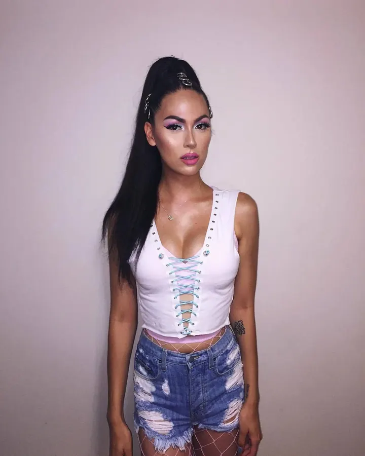 Femme trans européenne aux cheveux bruns-noirs, en haut noir, posant devant un fond blanc, avec des lunettes noires et des piercings visibles
