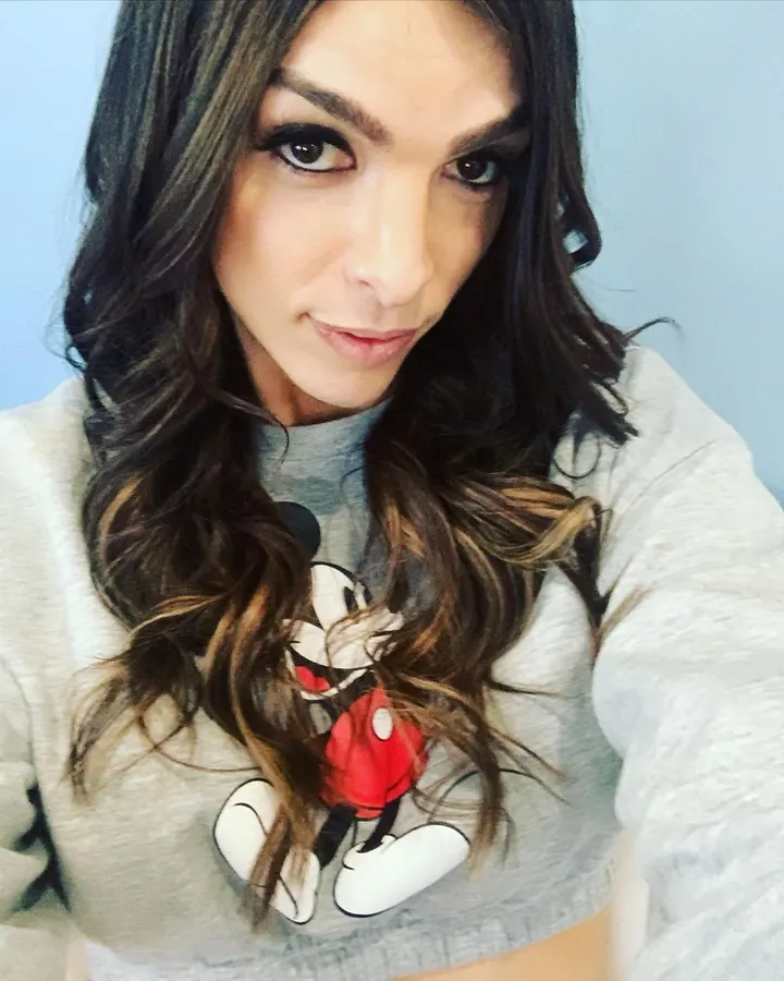 Femme trans européenne aux cheveux bruns, en sweat gris imprimé Mickey Mouse, regardant l'objectif
