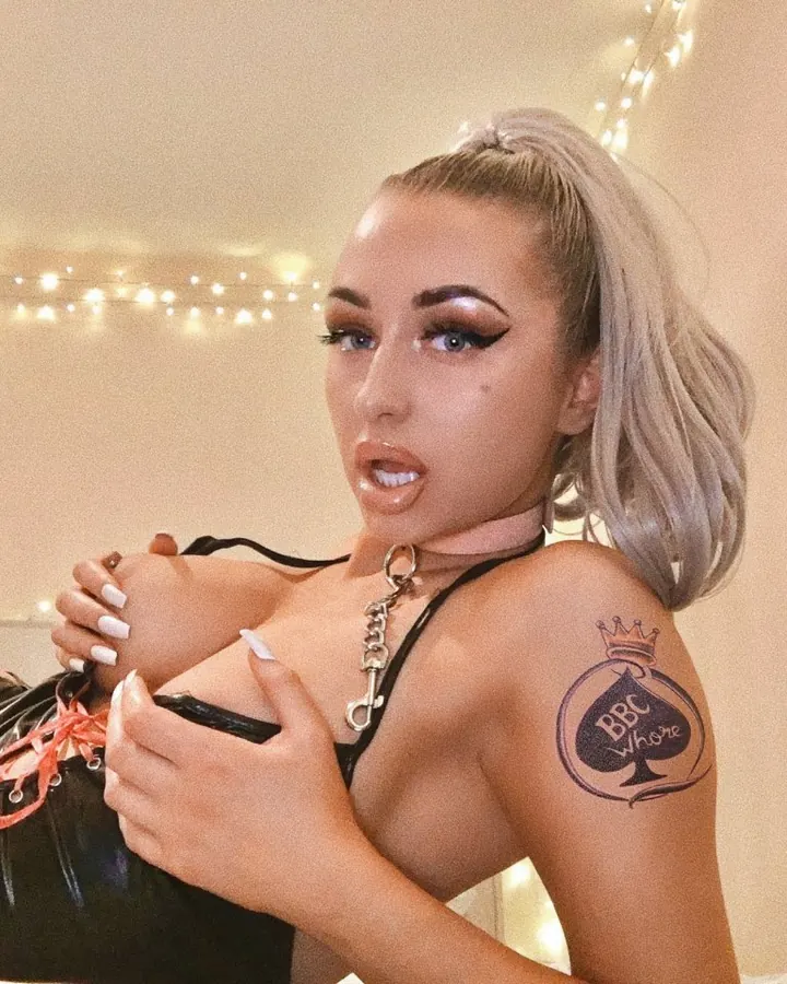 Femme trans aux cheveux platine, regardant l'appareil photo, portant un collier fin et une tenue sombre, dans un intérieur sobre.