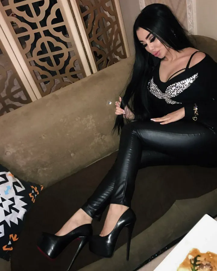 Femme trans aux cheveux bruns-noirs, regardant l'appareil photo, portant un collier fin et une tenue sombre, dans un intérieur sobre.