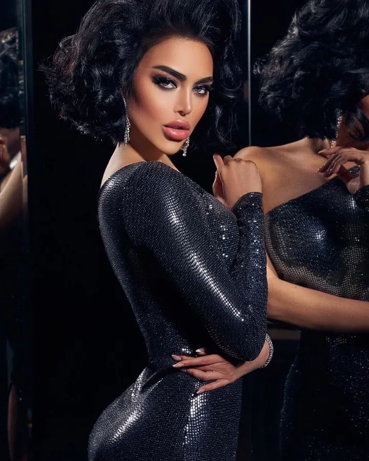 Femme trans aux cheveux bruns-noirs, regardant l'appareil photo, portant un collier fin et une tenue sombre, dans un intérieur sobre.