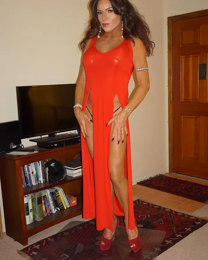 Femme trans en robe rouge longue fendue, posant dans un salon, mains sur les hanches