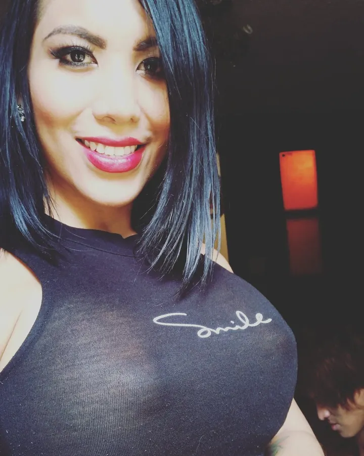 Femme trans souriante, en haut noir transparent, cheveux bleus, regardant l'appareil