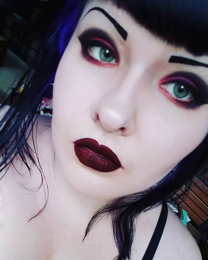 Femme trans aux cheveux noirs longs, portant un haut blanc, posant sur un lit, regardant l'appareil avec un sourire.
