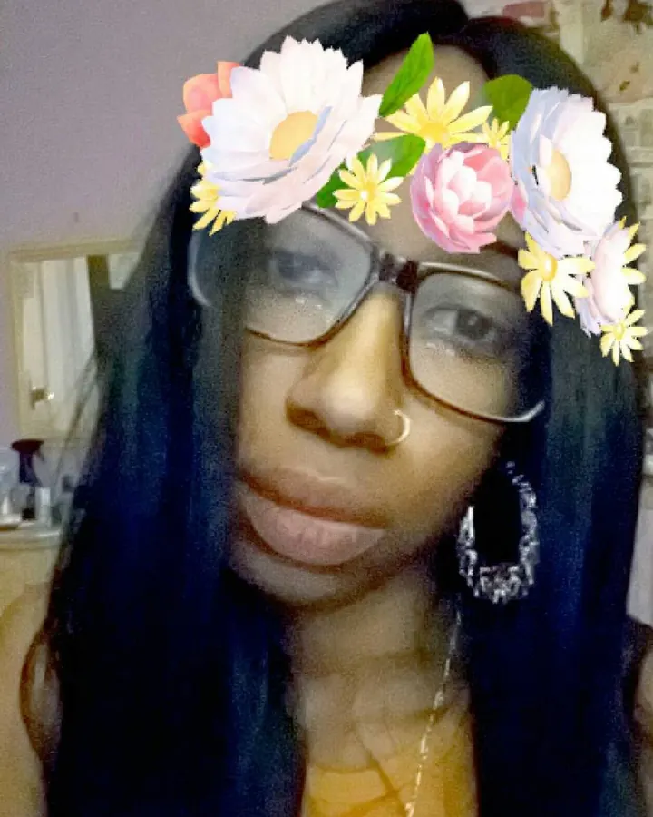 Femme trans aux cheveux noirs longs, portant des lunettes et un filtre floral, souriant doucement, dans un intérieur.
