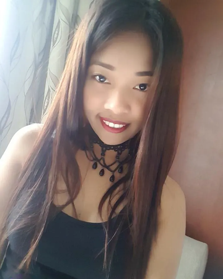 Femme trans aux cheveux châtains longs, portant un haut noir, posant sur un lit, regardant l'appareil avec un sourire.