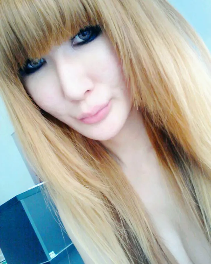 Femme trans aux cheveux blonds longs, regardant directement l'appareil, maquillage prononcé, dans un intérieur.