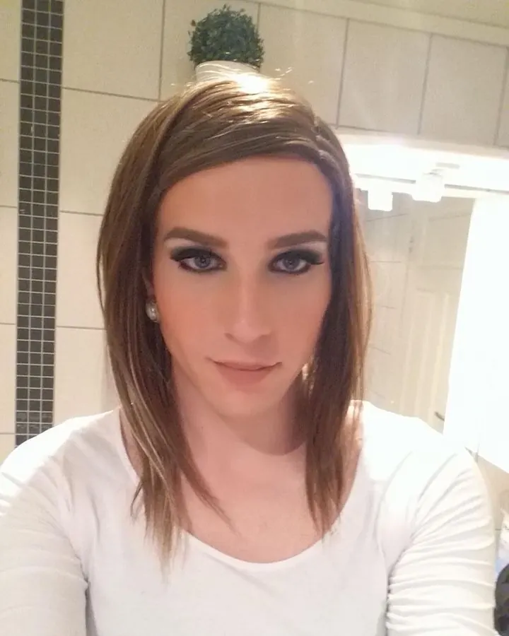 Femme trans en haut blanc, assise sur un lit, bras levé, tatouages visibles, chat blanc dormant à côté
