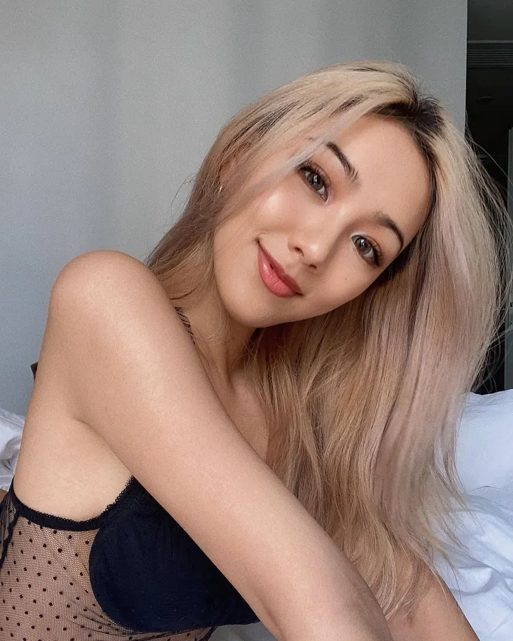 Femme trans européenne aux cheveux blonds et yeux noirs, en lingerie noire, allongée sur un lit, avec une lumière bleue en arrière-plan.