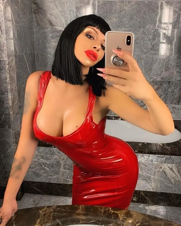 Femme trans aux cheveux noirs courts en robe rouge brillante, prenant un selfie dans un miroir devant un mur en marbre gris.