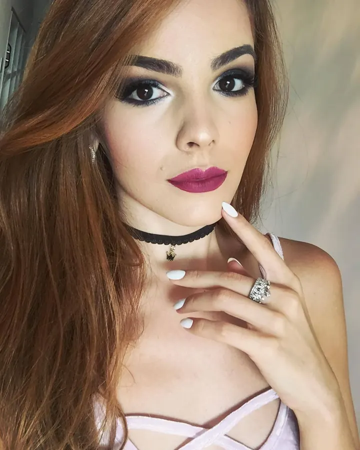 Jeune femme trans aux cheveux châtains, maquillée, portant un haut rose à lacets.