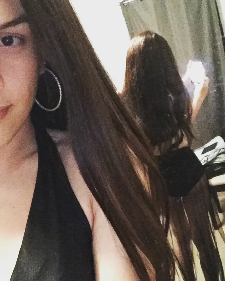 Femme trans aux cheveux bruns, prenant un selfie devant un miroir, portant un haut noir.