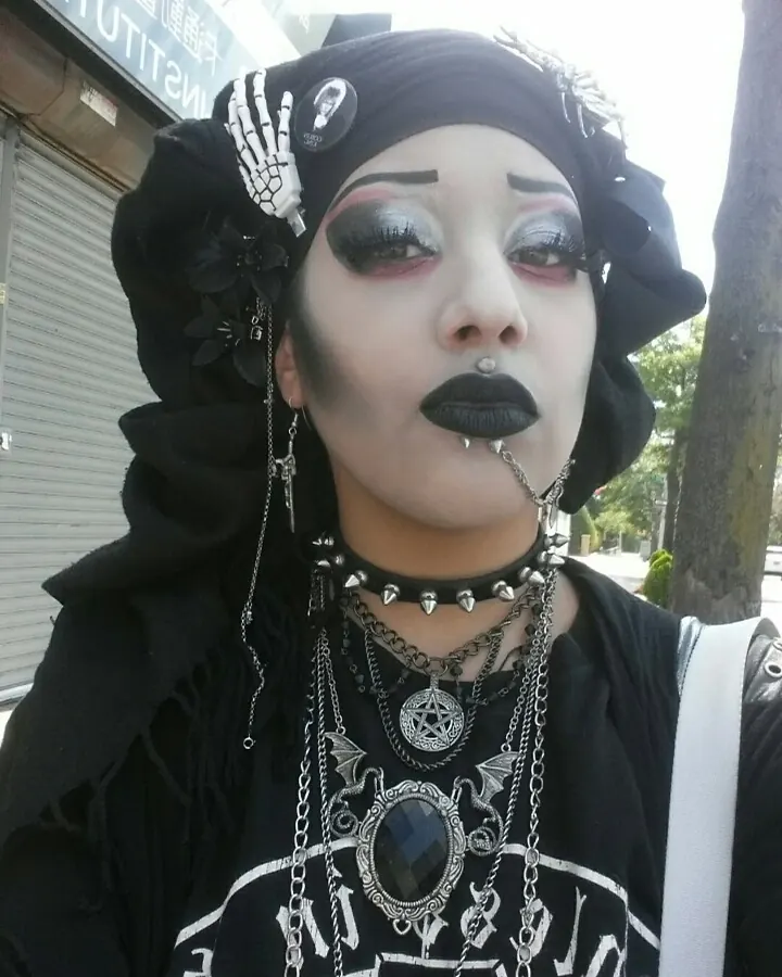 Femme trans aux cheveux noirs, portant un costume gothique noir avec des bijoux métalliques, posant dehors.