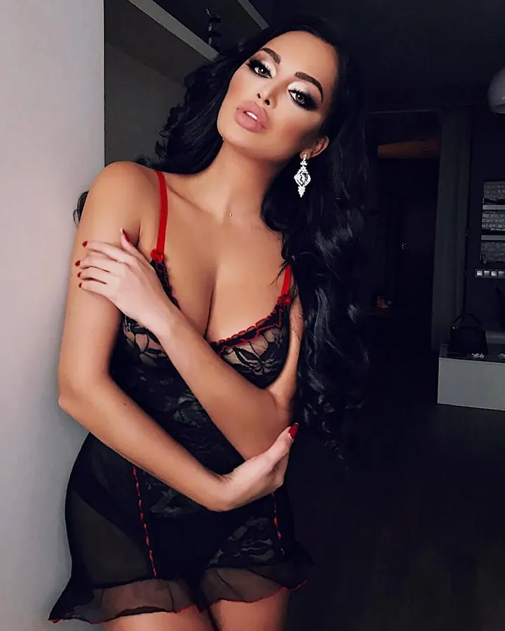 Femme trans aux cheveux noirs, portant un haut noir, posant avec un regard intense et un sourire discret.