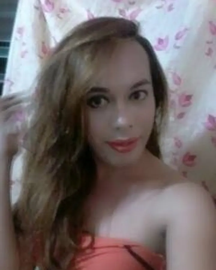 Femme trans aux cheveux châtains, portant un haut noir, posant avec un regard intense et un sourire discret.