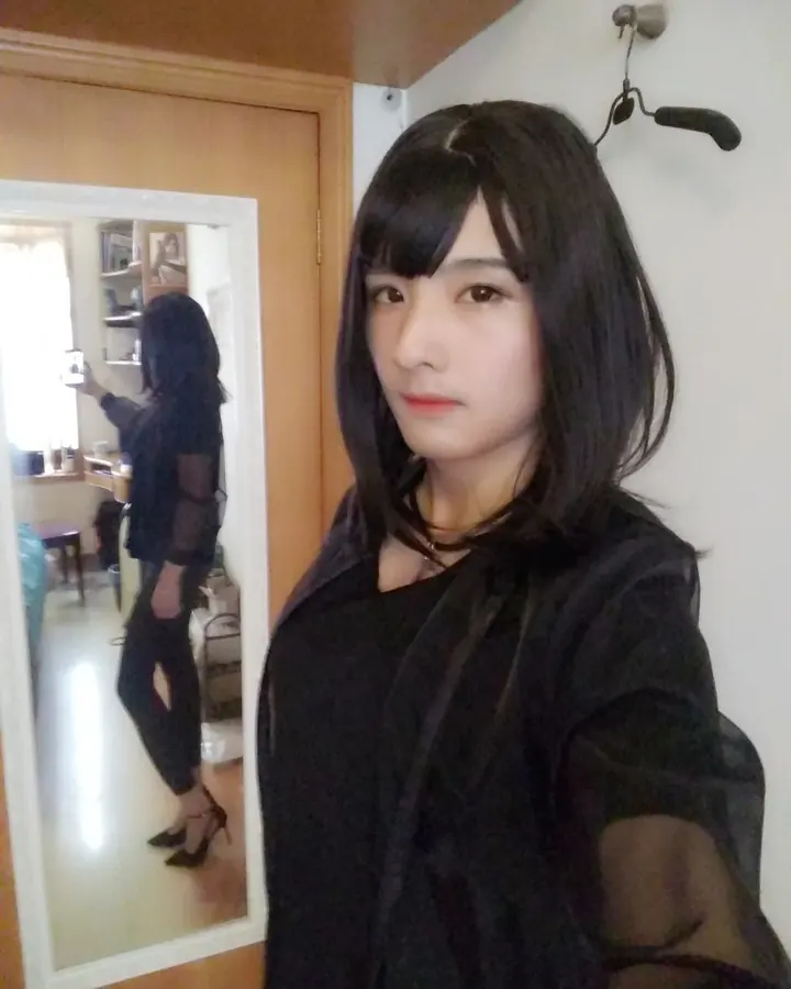 Jeune femme trans aux cheveux noirs, vêtue d’un haut noir transparent, prenant un selfie devant un miroir dans une pièce lumineuse.