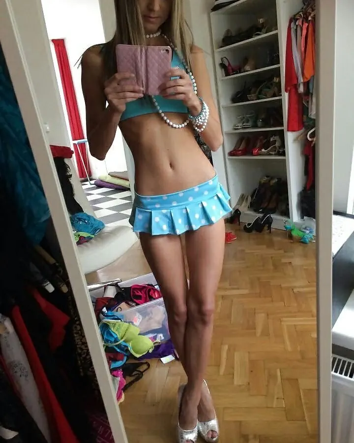 Femme trans aux cheveux blonds mi-longs, maquillage naturel, portant un haut noir et un bas noir, posant sur un lit.