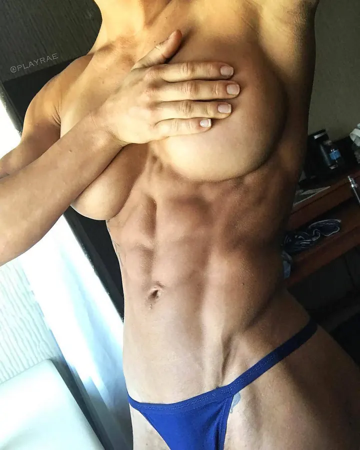 Femme trans aux cheveux blonds mi-longs, maquillage naturel, portant un haut noir et un bas noir, posant sur un lit.