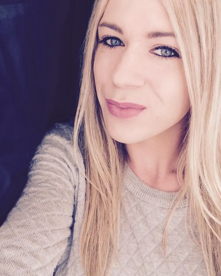 Femme trans aux cheveux blonds platine, maquillage naturel, portant un haut noir et un bas noir, posant sur un lit.